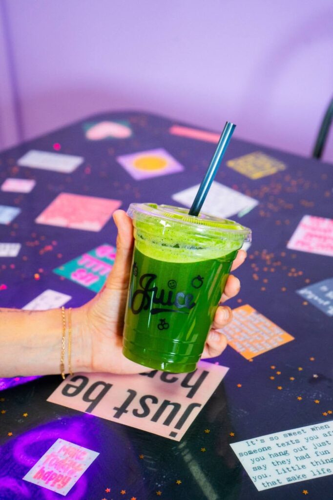 
A hand holds up a cup of vibrant green juice with a blue straw over a dark table decorated with various colorful notes and inspirational quotes.
