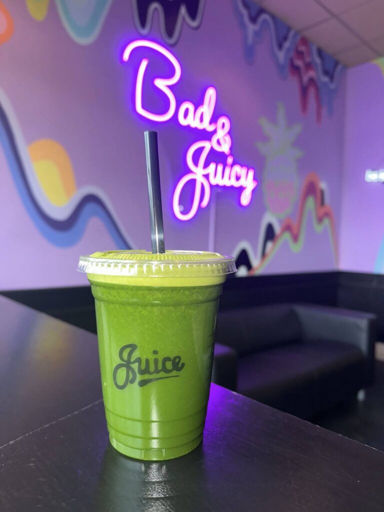A plastic cup of green juice sits on a table in front of a purple wall mural that features a bright purple neon sign reading "Bad & Juicy."